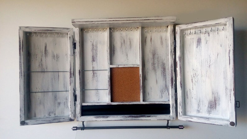 May include: White wooden jewelry cabinet with two doors open, revealing compartments for storing necklaces, earrings, and other jewelry. The cabinet has a distressed finish and a black metal rod for hanging necklaces.