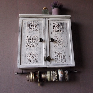 May include: White wooden cabinet with two doors and carved details. The cabinet has two brass knobs and a metal bar with hanging bracelets.
