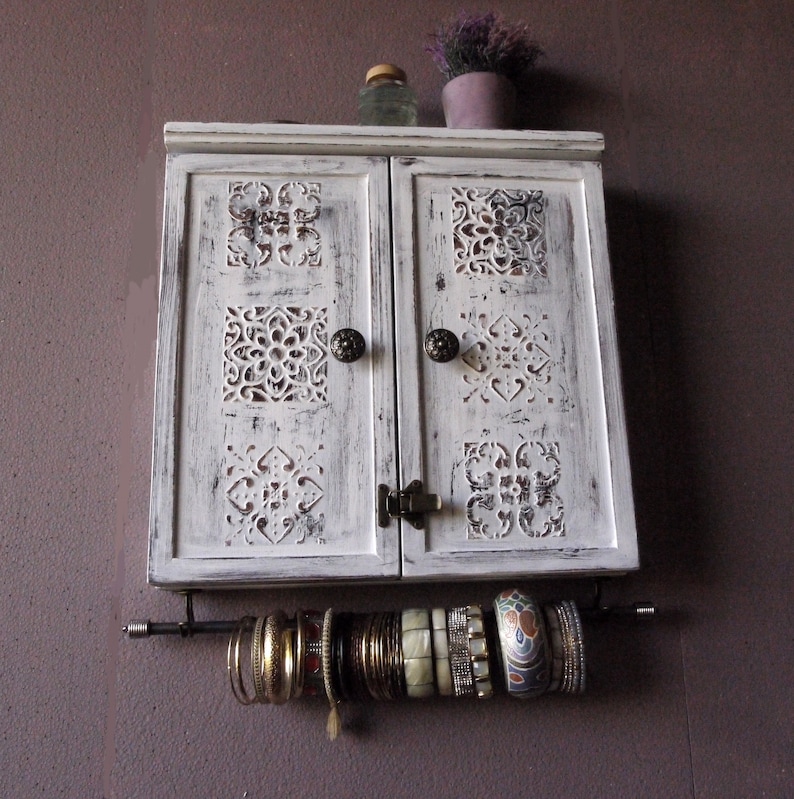 May include: White wooden cabinet with two doors and carved details. The cabinet has two brass knobs and a metal bar with hanging bracelets.