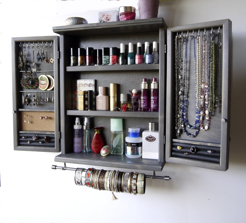 May include: A gray wooden jewelry cabinet with a mirrored door, three shelves, and a pull-out rod for hanging bracelets. The cabinet has a compartment for earrings and a section for necklaces.