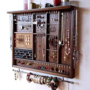 Può includere: Un organizer per gioielli in legno scuro con più scomparti e un'asta per braccialetti. L'organizer espone varie collane, orecchini e anelli. I gioielli sono in vari colori, tra cui argento, oro, rosso e verde.