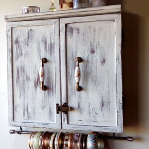 May include: A white distressed wooden cabinet with two doors and brass handles. The cabinet has a metal rod underneath for hanging jewelry. The rod is holding a variety of gold and silver bangles.