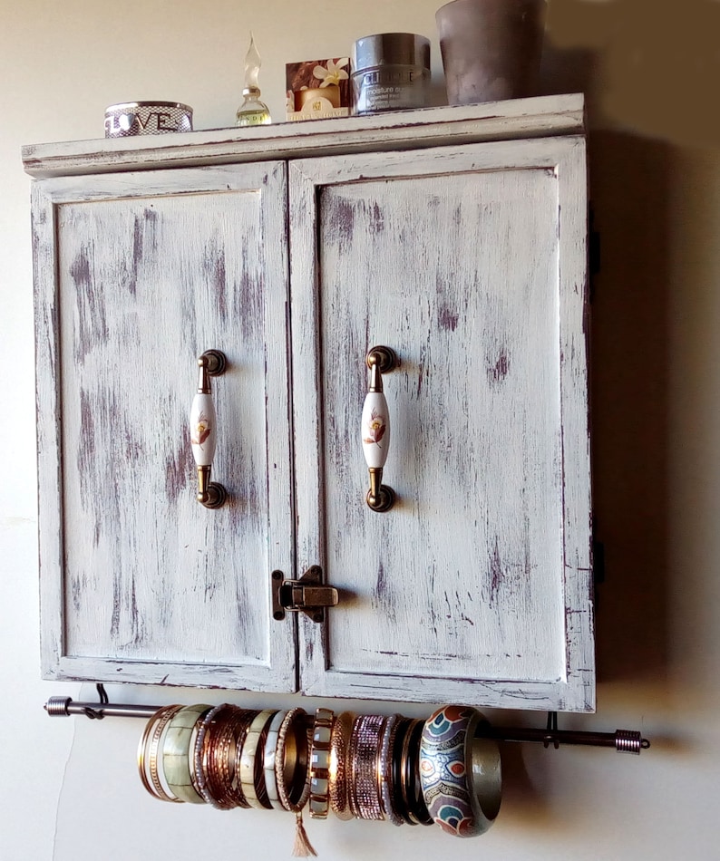 May include: A white distressed wooden cabinet with two doors and brass handles. The cabinet has a metal rod underneath for hanging jewelry. The rod is holding a variety of gold and silver bangles.