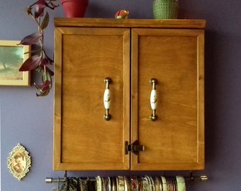 May include: A wooden cabinet with two doors and two brass knobs. The cabinet has a shelf on the inside and is mounted on the wall. There are several bracelets hanging from the bottom of the cabinet.