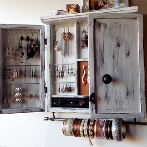 May include: White wooden jewelry cabinet with a distressed finish. The cabinet has two doors that open to reveal hooks for earrings and a small shelf for other jewelry. The cabinet also has a metal bar for hanging bracelets.