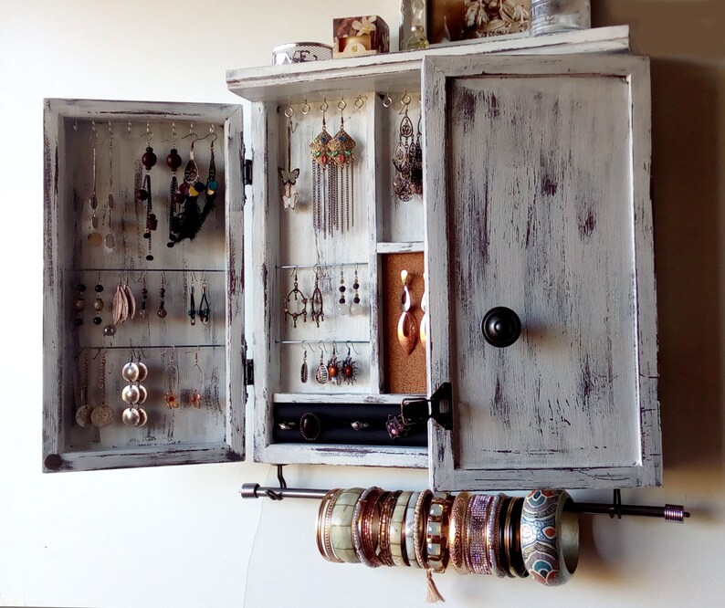 May include: White wooden jewelry cabinet with a distressed finish. The cabinet has two doors that open to reveal hooks for earrings and a small shelf for other jewelry. The cabinet also has a metal bar for hanging bracelets.