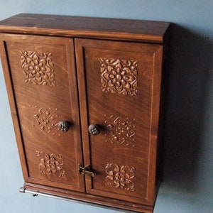 May include: A dark brown wooden cabinet with two doors, each door featuring a carved floral design. The cabinet has a metal bar underneath for hanging items.