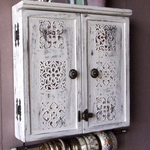 May include: White wooden cabinet with carved details and two doors. The cabinet has a metal rod underneath for hanging jewelry. The cabinet is mounted on a wall.