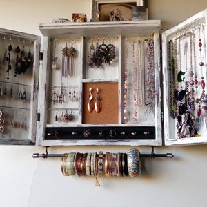 May include: White wooden jewelry organizer with two doors that open to reveal compartments for earrings, necklaces, and bracelets. The organizer has a corkboard section and a black velvet-lined section for rings.