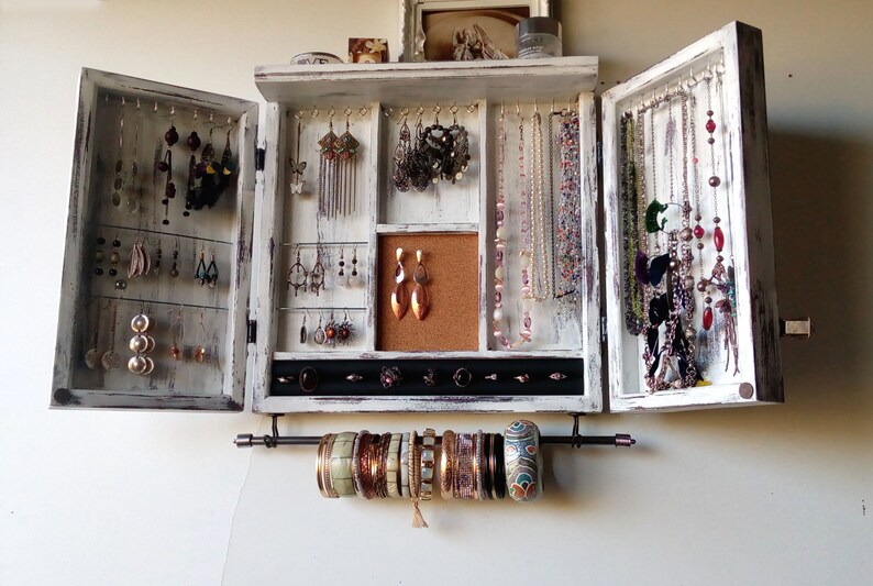 May include: White wooden jewelry organizer with two doors that open to reveal compartments for earrings, necklaces, and bracelets. The organizer has a corkboard section and a black velvet-lined section for rings.