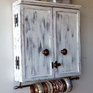 May include: White distressed wooden cabinet with two doors and brass hardware. A metal rod is attached to the bottom of the cabinet with a variety of bracelets hanging from it.