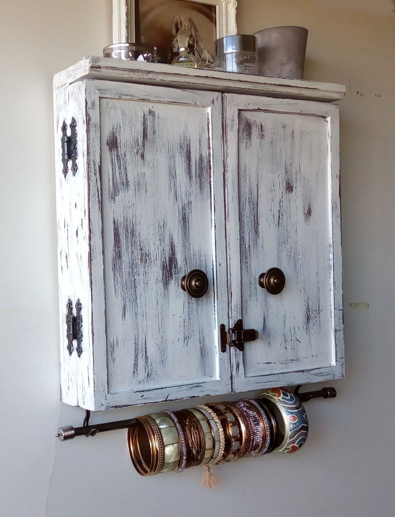 May include: White distressed wooden cabinet with two doors and brass hardware. A metal rod is attached to the bottom of the cabinet with a variety of bracelets hanging from it.
