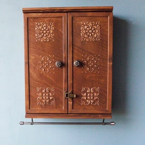 May include: A dark brown wooden cabinet with two doors and a metal towel bar. The doors have intricate carved designs and brass knobs.