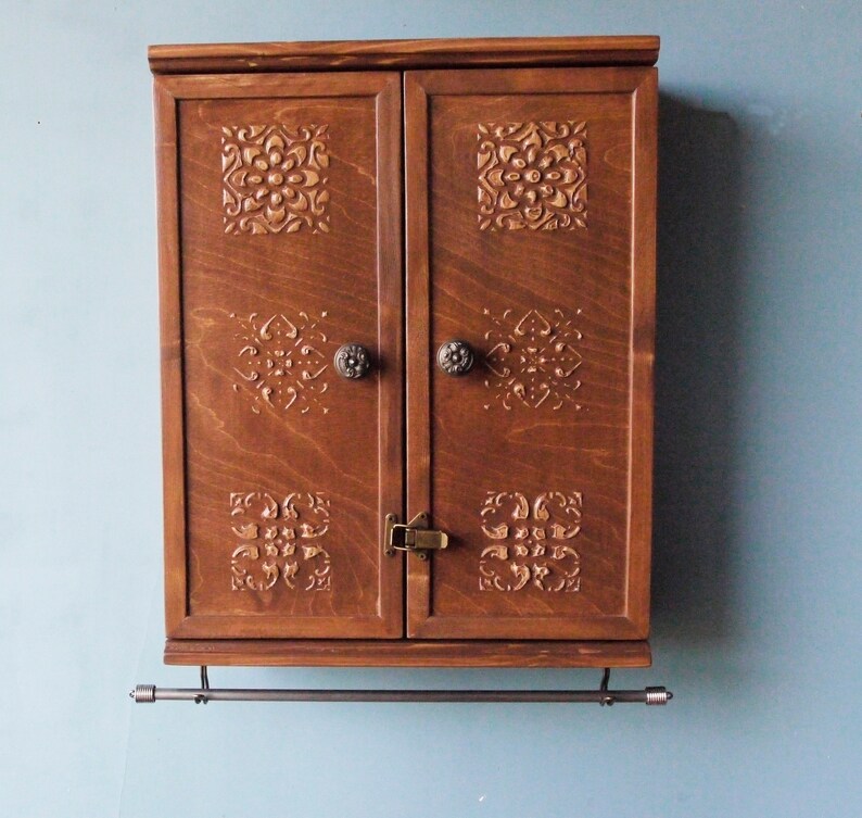 May include: A dark brown wooden cabinet with two doors and a metal towel bar. The doors have intricate carved designs and brass knobs.