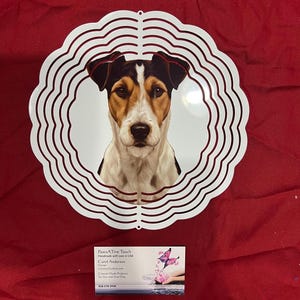May include: A circular white metal wind spinner featuring a detailed portrait of a tricolor dog with brown, black, and white fur. The spinner has a scalloped edge design and a glossy finish. A business card with the text PawsATive Touch is included.