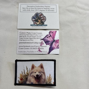 May include: A rectangular embroidery patch featuring a white dog with a pink tongue, surrounded by green and yellow foliage. Above it are two white business cards, one with a German Shepherd paw print logo and one with a purple butterfly graphic.