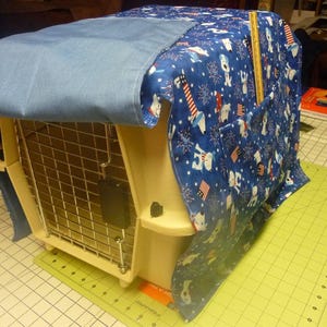 May include: A beige wire crate with a blue and white patterned fabric cover. The fabric has a design of white dogs and fireworks on a blue background.