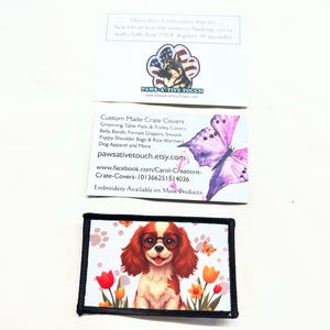 May include: A rectangular embroidered patch featuring a brown and white spaniel dog wearing round glasses, surrounded by colorful tulips and paw prints. Above the patch are two white business cards with text and graphics including a dog and a butterfly.