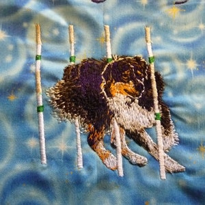 May include: Embroidered fabric featuring a dog in agility training, jumping over poles. The dog has black, brown, and white fur. The background is a light blue fabric with a starry pattern. The poles are white with green and tan accents.