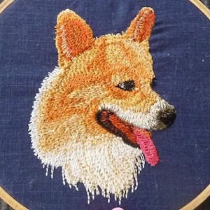 May include: Embroidered portrait of a dog's head on a blue fabric background. The dog has orange and white fur, a black nose, and a pink tongue sticking out. The dog's ears are perked up.