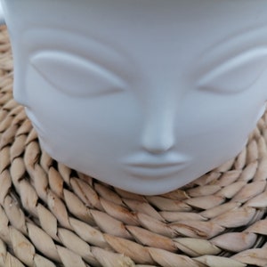 Może przedstawiać: Biała ceramiczna doniczka w kształcie twarzy z zamkniętymi oczami i neutralnym wyrazem. Doniczka stoi na plecionym słomianym dywaniku.