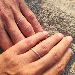 Könnte beinhalten: Zwei Hände, eine männliche und eine weibliche, mit passenden Silberringen. Die Ringe sind dünne Metallbänder und werden an den Ringfingern getragen. Der Hintergrund ist eine hellgraue Steinoberfläche.