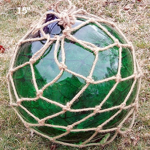 Può includere: Un galleggiante da pesca in vetro verde, di circa 38 cm di diametro, racchiuso in una rete di corda beige. Il galleggiante ha una parte superiore blu scuro. La rete di corda è annodata attorno alla sfera di vetro.