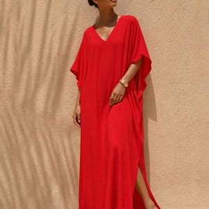 Può includere: Un vivace abito caftano rosso, lungo fino al pavimento, con scollo a V e maniche corte. L'abito presenta uno spacco laterale alto, offrendo una silhouette fluida. La modella indossa un braccialetto dorato. Lo sfondo è un muro beige neutro con ombre.