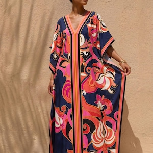 May include: A flowing, V-neck caftan dress with a vibrant floral pattern in shades of pink, orange, and white against a navy blue background. The dress features a bold black and orange stripe down the center and along the edges.