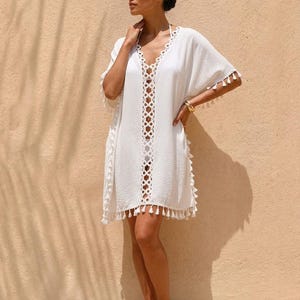 Könnte beinhalten: Weißes Strandkleid mit V-Ausschnitt und Lochmuster auf der Vorderseite. Das Kleid hat kurze Ärmel und einen Fransenbesatz. Das Model trägt ein goldenes Armband. Der Hintergrund ist eine hellbraune Wand.