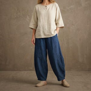 Könnte beinhalten: Ein beiges Leinenoberteil mit Dreiviertelärmeln und einem einzelnen Knopfverschluss, kombiniert mit einer locker sitzenden, dunkelblauen Leinenhose. Die Hose hat ein weites Bein und eine entspannte Passform. Das Outfit ist lässig und bequem.