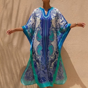 Puede incluir: Un caftán largo y fluido en tonos azules, turquesas y blancos. La prenda presenta un cuello en V y mangas anchas, adornada con un patrón ornamental y arremolinado. El diseño del caftán incluye un panel vertical central y ribetes turquesa.