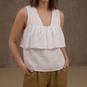 Peut inclure: Un haut blanc sans manches avec un col en V et des volants superposés. Le haut est associé à un pantalon vert olive. La tenue est faite d'un tissu léger, idéal pour le temps chaud. Le modèle a les mains dans les poches.