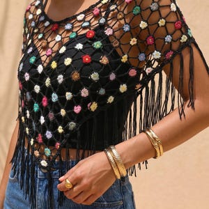 May include: Black crochet top with colorful floral accents and fringe detailing. The top is worn over a black tank top and paired with blue jeans. Gold bracelets and a ring complete the look.