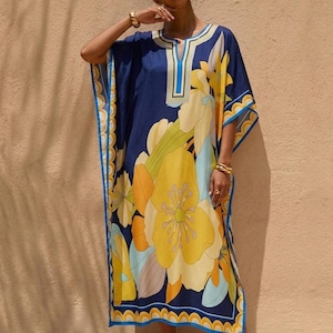 Puede incluir: Un vestido caftán azul marino con un gran estampado floral en amarillo, naranja y azul claro. El vestido tiene un escote en V con ribete azul y amarillo y un borde festoneado. El vestido llega a la rodilla.
