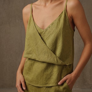Könnte beinhalten: Olivgrünes Camisole-Top mit einem geschichteten Design. Das Top hat dünne Spaghettiträger und eine Wickeloptik vorne. Der Stoff scheint ein leichter Leinenstoff oder ähnliches zu sein, mit einer subtilen Textur. Der Stil ist lässig und bequem.