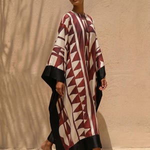 May include: A flowing, floor-length caftan in cream, burgundy, and black. The caftan features a geometric pattern of burgundy triangles and vertical stripes. The sleeves and hem are trimmed with black fabric. The model is barefoot.