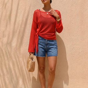 May include: A woman wearing a red long-sleeve top tied at the waist, paired with blue denim shorts. She holds a small woven handbag. The background is a light peach color with shadows.