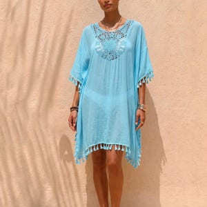 Könnte beinhalten: Ein hellblauer Kaftan mit einem runden Häkeldetail am Ausschnitt und kurzen Ärmeln. Das Kleidungsstück ist mit einer Reihe kleiner, passender Quasten am Saum und an den Ärmelkanten versehen, was für ein strukturiertes und leichtes Aussehen sorgt.