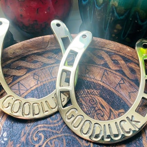 May include: Two gold-colored horseshoes with the words "Good Luck" engraved on them. The horseshoes are on a wooden surface with a decorative pattern.