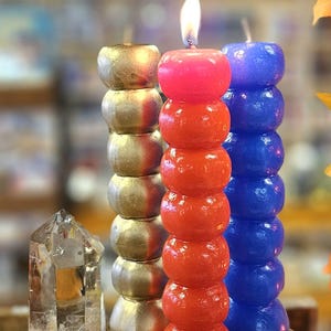 May include: Three pillar candles, one gold, one red, and one blue. Each candle is made of multiple stacked spheres. The red candle is lit.