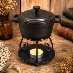 May include: A black cast iron pot with a matching lid and side handles, resting on a black metal stand with a circular base. A small, lit tea light candle sits on the base of the stand, positioned directly beneath the center of the pot.