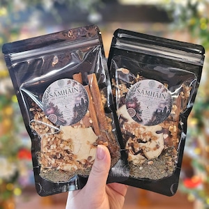 May include: Two black resealable pouches containing Samhain Simmer Pot ingredients. Each label features the text Samhain Simmer Pot, listing apple, rosemary, cinnamon, clove, and orange peel. The contents include dried fruit slices, cinnamon sticks, and herbs.