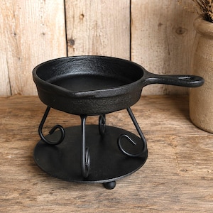 May include: A black cast iron mini skillet resting on a black metal stand with three curved legs and a circular base. The skillet features a short handle and a textured surface, while the stand provides a stable elevated platform for the pan.