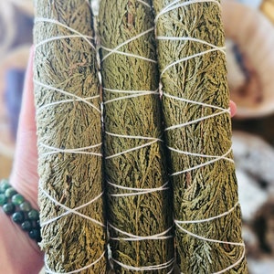 May include: Three bundles of dried cedar smudge sticks, tied with twine, are held in a hand. The sticks are a light green color and have a rough texture.