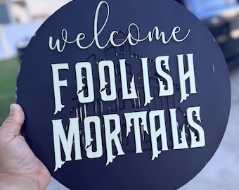 Könnte beinhalten: Runder, marineblauer Holzschild mit den Worten "welcome FOOLISH MORTALS" in weißer, mehrschichtiger Schrift. Das Schild hat einen Used-Look und eine Gebäudesilhouette.