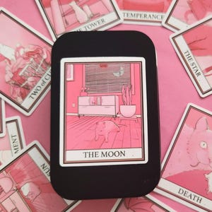 May include: A pink tarot card with a black border and the text "THE MOON" in white. The card depicts a pink room with a cat in the centre, a dresser, and a window with blinds.