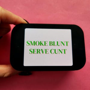 Könnte beinhalten: Ein schwarzes, rechteckiges Objekt mit abgerundeten Ecken, das den grünen Text "SMOKE BLUNT SERVE CUNT" auf weißem Grund zeigt. Das Objekt wird vor einem rosa Hintergrund gehalten.