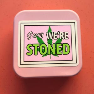 May include: A pale pink square container with rounded edges. The container features a sticker with the text "Sorry We're Stoned" in white and green, with a green cannabis leaf on a pink background.