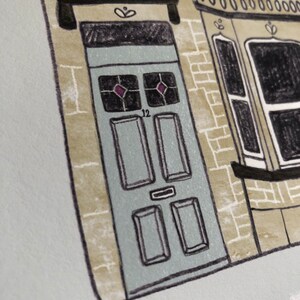 Op de afbeelding: Een handgetekend ontwerp van een huis met een blauwe deur. De deur heeft het nummer "12" erop en twee ruitvormige ramen. Het huis is van baksteen en heeft witte versieringen.